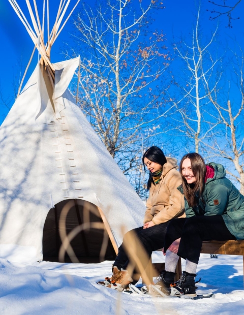 Two women sitting outside on a bench in the wintertime, with a teepee in the background